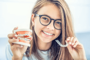 young girl with glasses holding a 4D model of teeth with braces in one hand and Invisalign aligners in the other cosmetic dentistry dentist in Towson Maryland